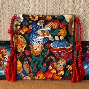 May include: A black drawstring bag with a colorful pattern of dolls, apples, and autumn leaves. The bag has red rope drawstrings with tassels. Two tarot cards, "Coins" and "The Fool", are placed in front of the bag.