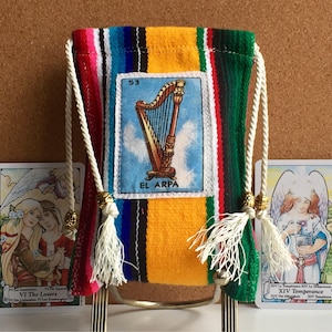 May include: A colorful, rectangular drawstring bag with a woven striped pattern in red, yellow, green, and blue. The bag features a card with a harp illustration and the text "EL ARPA". Two tarot cards flank the bag.