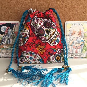 May include: A red drawstring bag featuring a sugar skull design, with blue rope ties and tassels. Two tarot cards are positioned on either side of the bag, one showing Death and the other Temperance. The bag is suitable for tarot cards.