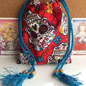 May include: A red drawstring bag with a sugar skull pattern, blue rope handles, and tassels. The bag features a repeating design of white sugar skulls with floral accents. Gold beads adorn the rope where the tassels are attached. Tarot cards are visible in the background.