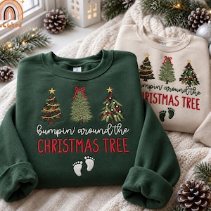 May include: A dark green sweatshirt and a beige sweatshirt, both featuring embroidered Christmas trees, the text "bumpin' around the CHRISTMAS TREE" in white and red, and a pair of small white baby footprints at the bottom of the design.