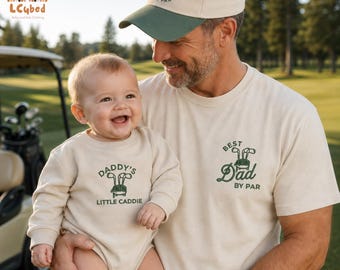 May include: A beige t-shirt and cap set with the text "Best Dad by Par" and a golf club graphic. A baby in a beige onesie with "Daddy's Little Caddie" text and golf club graphic is held by an adult.