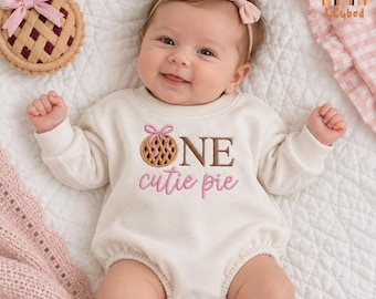 May include: A cream-colored long-sleeve baby romper featuring an embroidered graphic of a lattice-top pie with a pink bow and the text "ONE cutie pie" in brown and pink lettering. The baby wears a matching cream headband with a pink bow.