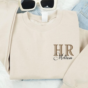 May include: A beige sweatshirt with the embroidered letters "HR" and the name "Melissa" in a script font. The sweatshirt is paired with white sunglasses and blue jeans. Black and white sneakers are visible at the bottom.