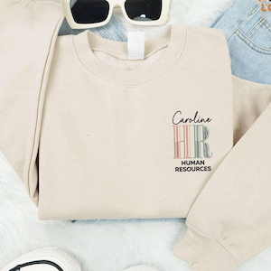 May include: A beige crewneck sweatshirt with the text "Caroline HR HUMAN RESOURCES" in a colorful design. The sweatshirt is paired with white sunglasses and black and white sneakers.