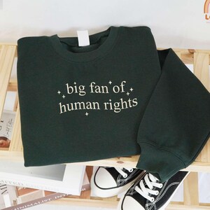 May include: A dark green sweatshirt with the text "big fan of human rights" in white script. The sweatshirt is folded on a wooden shelf, with a pair of black and white sneakers visible below. The sweatshirt has a crew neck and long sleeves.