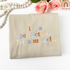 May include: A beige sweatshirt with the embroidered phrase "it's a perfect day for a miracle" in colorful thread. The sweatshirt is folded and displayed with decorative beads and pink roses.