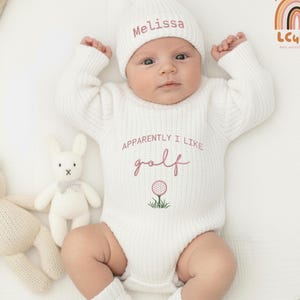 May include: A baby wearing a white knit outfit, including a hat embroidered with "Melissa" and a onesie with "Apparently I like golf" and a golf ball design. A teddy bear and a bunny toy are nearby.