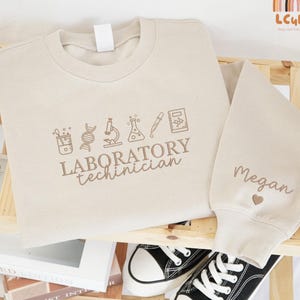 May include: A beige sweatshirt and matching sleeve with the name "Megan" embroidered on the sleeve. The sweatshirt features laboratory-themed graphics and the words "Laboratory Technician". Black and white sneakers are visible.