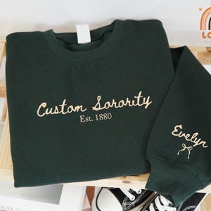 May include: A dark green sweatshirt with the words "Custom Sorority Est. 1880" embroidered in a light tan color. The sleeve has the name "Evelyn" embroidered in the same color, with a bow. Black and white sneakers are visible.