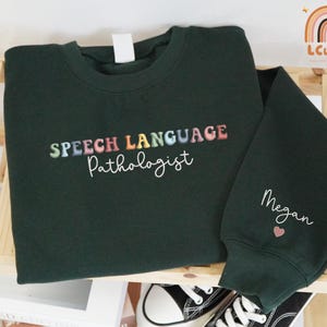 May include: A dark green sweatshirt with the words "SPEECH LANGUAGE Pathologist" in colorful embroidery. The sleeve has the name "Megan" embroidered in white with a small pink heart. Black and white sneakers are visible.