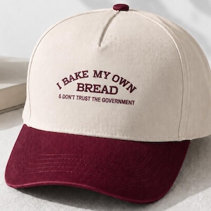 May include: A two-tone baseball cap with a cream-colored crown and a dark maroon brim. The front of the crown features embroidered dark maroon text that reads I BAKE MY OWN BREAD & DON'T TRUST THE GOVERNMENT in a curved, bold, sans-serif font.