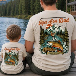 May include: Beige t-shirts with fishing-themed graphics. One shirt reads "Reel Cool Dad" with a mountain and fish design, the other reads "Reel Cool Kid". Both shirts feature a fisherman, fish, and the words "Hooked on Adventure".