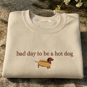 May include: Beige sweatshirt with embroidered text "bad day to be a hot dog" and a dachshund in a hot dog bun. The sweatshirt is folded and rests on a textured, gray surface. The design is in brown thread.