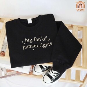 May include: Black sweatshirt with the text "big fan of human rights" in a white, stylized font. The sweatshirt is folded on a wooden shelf, with a pair of black and white sneakers visible below. The overall aesthetic is casual and modern.