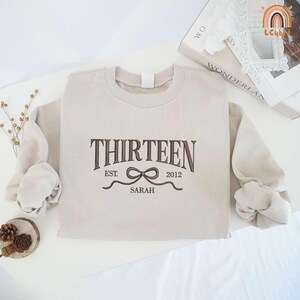 May include: A beige sweatshirt with the text "THIRTEEN EST. 2012 SARAH" and a brown ribbon design.