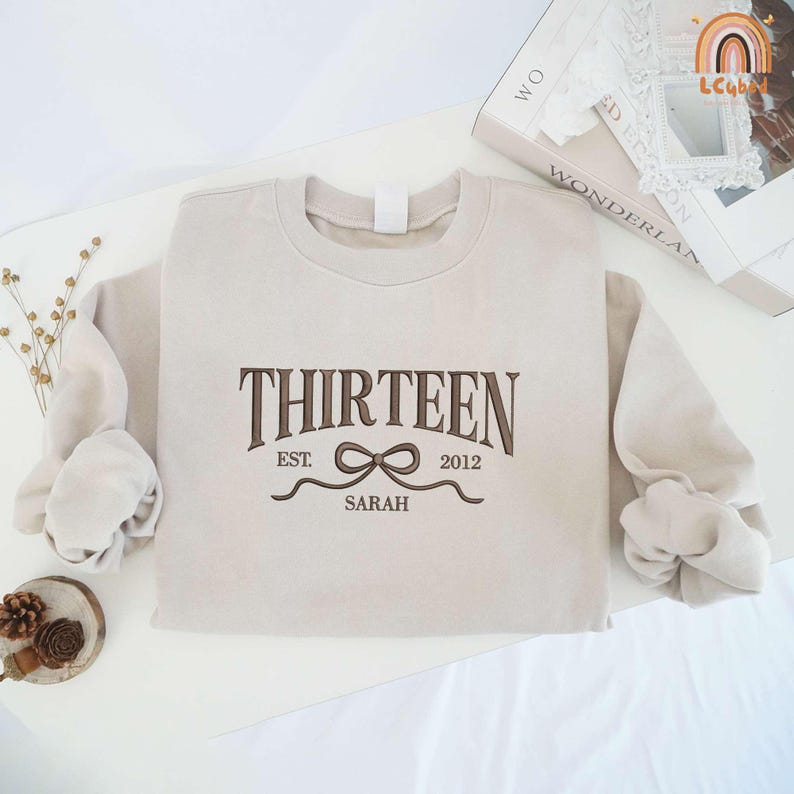 May include: A beige sweatshirt with the text "THIRTEEN EST. 2012 SARAH" and a brown ribbon design.