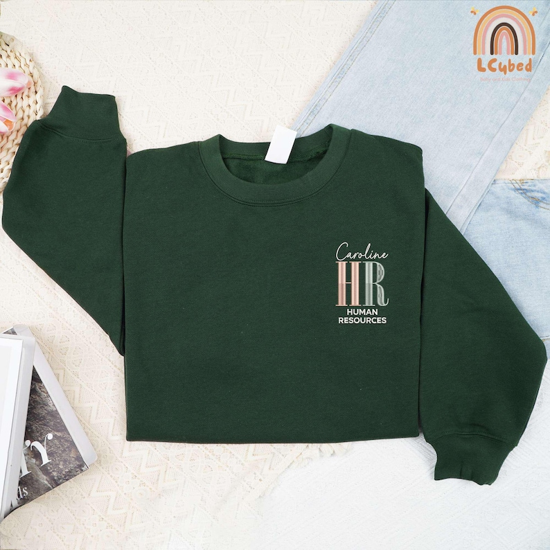 May include: A dark green sweatshirt with the text "Caroline HR HUMAN RESOURCES" in a stylish font. The letters "HR" are in a vertical striped design with pastel colors. The sweatshirt is laid flat on a light-colored surface.