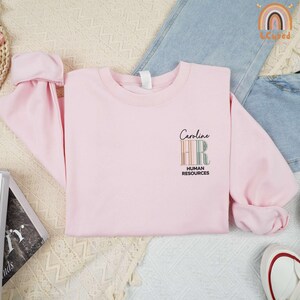 May include: A light pink sweatshirt with the text "Caroline HR HUMAN RESOURCES" in a colorful design. The long-sleeved top is folded on a cream-colored surface with a pair of blue jeans and a white shoe in the background.