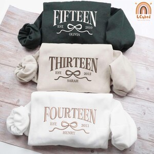May include: Three white sweatshirts with different numbers and names embroidered on them. The top sweatshirt says "FIFTEEN EST. 2010 OLIVIA", the middle sweatshirt says "THIRTEEN EST. 2012 SARAH", and the bottom sweatshirt says "FOURTEEN EST. 2011 HENRY".