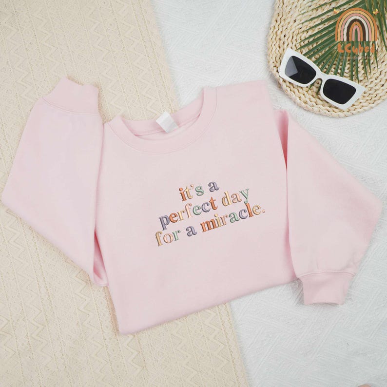 May include: A light pink sweatshirt with the embroidered text "it's a perfect day for a miracle" in colorful thread. The sweatshirt is displayed on a cream-colored surface.
