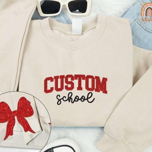 May include: A beige sweatshirt with the text "CUSTOM SCHOOL" in red glitter lettering. A red glitter bow is on the left sleeve.