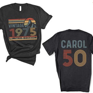 May include: Two black t-shirts with vintage-style graphics. The first shirt says "Vintage Est. 1975 Limited Edition" with a mountain and sunset graphic. The second shirt says "Carol 50" with a rainbow stripe graphic.