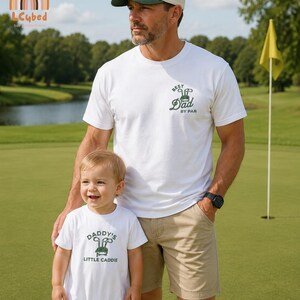 May include: A man and a child are on a golf course. The man wears a white t-shirt with "Best Dad by Par" and a beige cap. The child wears a white onesie with "Daddy's Little Caddie". The background includes a golf flag and a lake.