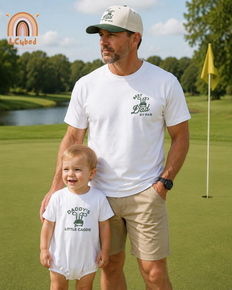 May include: A man and a child are on a golf course. The man wears a white t-shirt with "Best Dad by Par" and a beige cap. The child wears a white onesie with "Daddy's Little Caddie". The background includes a golf flag and a lake.