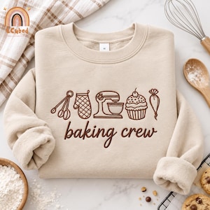 May include: A beige crewneck sweatshirt featuring brown embroidery of baking tools including measuring spoons, an oven mitt, a stand mixer, a cupcake, and a piping bag, with the text "baking crew" embroidered in a cursive font below the icons.
