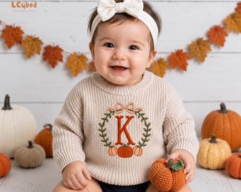 May include: A beige long-sleeve knit sweater featuring an embroidered design of a wreath made of green leaves and small orange pumpkins surrounding a large orange letter K, topped with a small bow. The sweater is worn with a white headband with a bow.