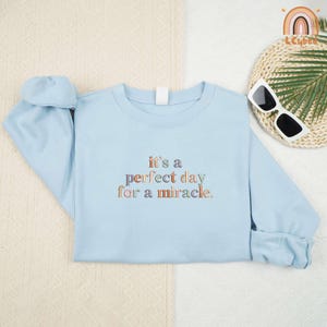 May include: A light blue sweatshirt with the embroidered text "it's a perfect day for a miracle" in colorful lettering. The long-sleeved top is folded on a cream-colored surface, with sunglasses and a decorative item in the background.