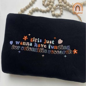 May include: Black sweatshirt with colorful embroidered text that reads "girls just wanna have funding for scientific research." The design includes stars and floral accents in orange, blue, and white.