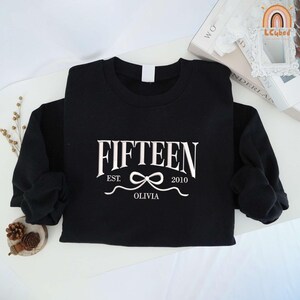May include: Black sweatshirt with white text that reads "FIFTEEN EST. 2010 OLIVIA" with a white bow in between the dates.
