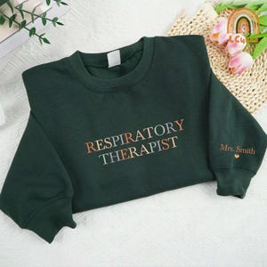 May include: A dark green sweatshirt with the text "RESPIRATORY THERAPIST" in rainbow colors. The text "Mrs. Smith" is on the right sleeve with a small heart.