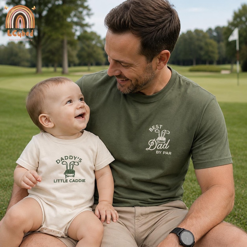 May include: A father and baby are smiling at each other while sitting on a green lawn. The father wears an olive green t-shirt with "Best Dad by Par" printed on it. The baby wears a beige onesie with "Daddy's Little Caddie" printed on it.