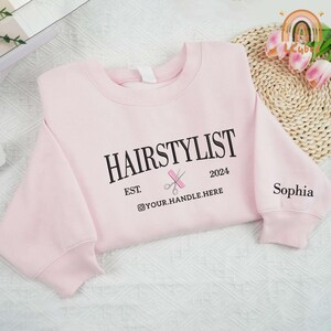 May include: Pink sweatshirt with the text "HAIRSTYLIST EST. 2024" and a pair of scissors and a comb graphic. The text "@YOUR.HANDLE.HERE" is below the graphic. The text "Sophia" is on the right sleeve.