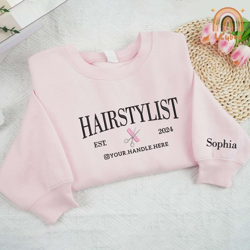 May include: Pink sweatshirt with the text "HAIRSTYLIST EST. 2024" and a pair of scissors and a comb graphic. The text "@YOUR.HANDLE.HERE" is below the graphic. The text "Sophia" is on the right sleeve.