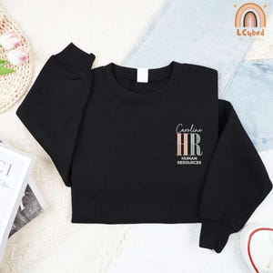 May include: Black crewneck sweatshirt with the text "Caroline HR HUMAN RESOURCES" in a script font and pastel colors. The sweatshirt is made of a soft material and has long sleeves.