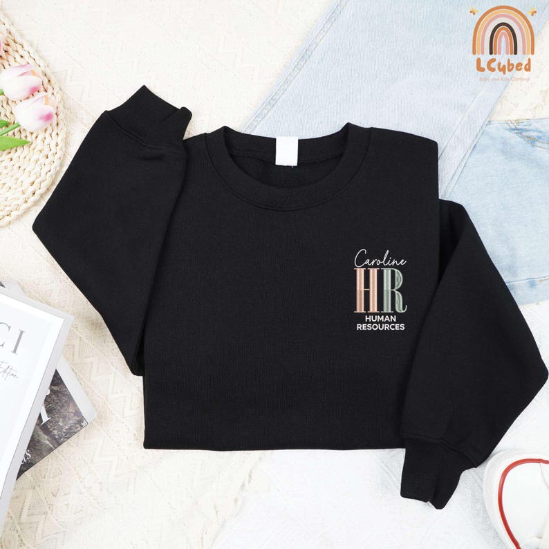 May include: Black crewneck sweatshirt with the text "Caroline HR HUMAN RESOURCES" in a script font and pastel colors. The sweatshirt is made of a soft material and has long sleeves.