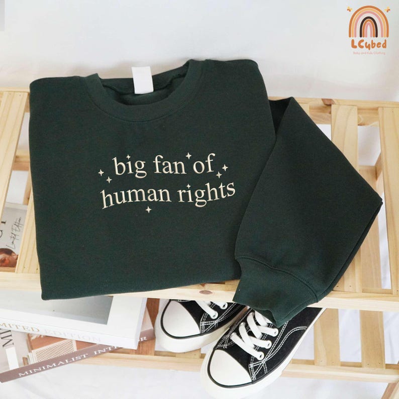May include: A dark green sweatshirt with the text "big fan of human rights" in white script. The sweatshirt is folded on a wooden shelf, with a pair of black and white sneakers visible below. The sweatshirt has a crew neck and long sleeves.