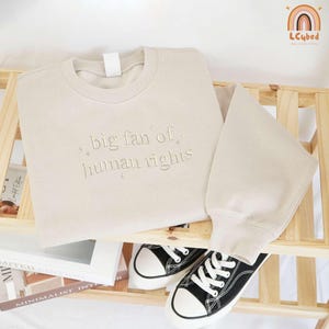 May include: A beige sweatshirt and matching pants set with the embroidered text "big fan of human rights." Black canvas sneakers with white soles and laces are placed below the set. The set is displayed on a wooden shelf.