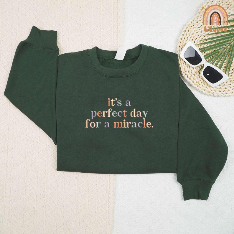 May include: A dark green sweatshirt with the phrase "it's a perfect day for a miracle" in a multi-colored font. The sweatshirt is folded on a white and beige textured surface. Sunglasses and a decorative item are in the background.