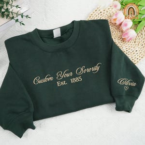 May include: Dark green sweatshirt with embroidered text "Custom Your Sorority Est. 1885" in a cursive font. The sleeve features the name "Olivia" and a heart symbol. The sweatshirt is made of a soft material.