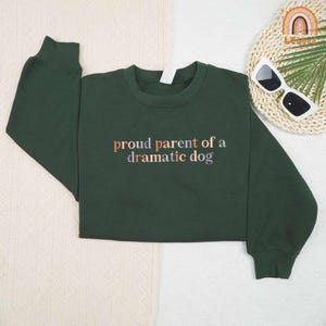 May include: A dark green sweatshirt with the text "proud parent of a dramatic dog" in a multi-colored font. The sweatshirt is folded on a white surface.