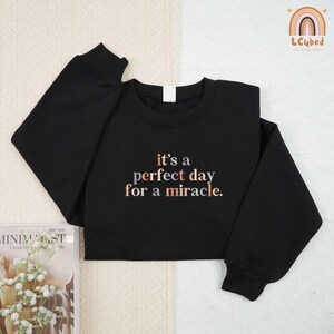 May include: Black sweatshirt with the text "it's a perfect day for a miracle" in colorful lettering. The sweatshirt has long sleeves and a crew neck. The brand logo is in the upper right corner.