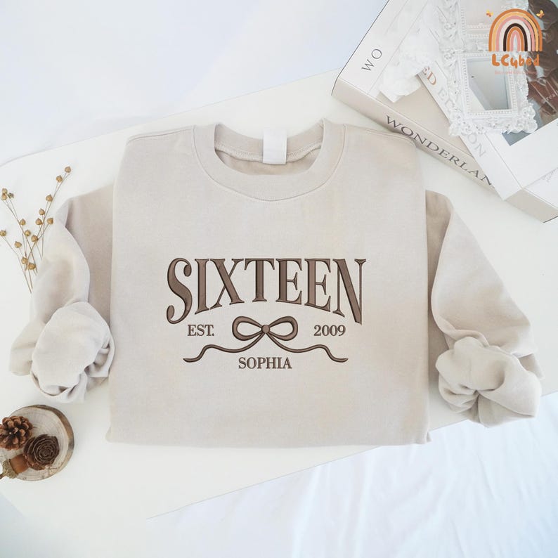 May include: A beige sweatshirt with long sleeves. The front of the sweatshirt features the word "SIXTEEN" in brown script, with "EST. 2009" and the name "SOPHIA" below. A bow graphic is centered under the word "SIXTEEN".