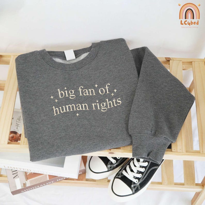 May include: A gray sweatshirt with the text "big fan of human rights" in white. The sweatshirt is displayed on a wooden shelf with a pair of black and white sneakers. The sweatshirt has a crew neck and long sleeves.