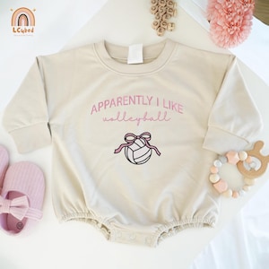 May include: A beige baby onesie with long sleeves, featuring the embroidered text "Apparently I like volleyball" in pink, along with a volleyball graphic. The onesie has a round neckline and snap closures.