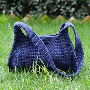 Può includere: Una borsa a tracolla in crochet blu scuro con un cinturino lungo. La borsa è appoggiata su un letto di erba verde.